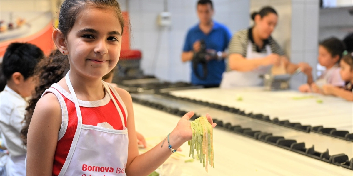 Üzerinde Bornova Belediyesi yazılı beyaz bir önlük giymiş küçük bir kız çocuğu, elinde tuttuğu taze yeşil ve sarı renkli makarna şeritlerini kameraya doğru göstererek gülümsüyor. Arka planda mutfak tezgahı başında makarna hazırlayan diğer çocuklar ve onlara yardımcı olan yetişkinler bulunuyor.