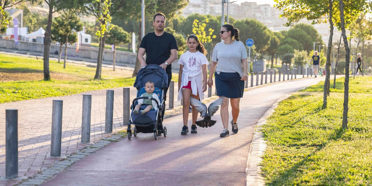 Güneşli bir günde parktaki yürüyüş yolunda ilerleyen bir aile; siyah tişörtlü bir erkek bebek arabasını sürerken, yanında beyaz tişörtlü genç bir kız ve gri tişörtlü bir kadın yürüyor. Bebek arabasında küçük bir çocuk otururken, önlerinde kanat çırpan bir güvercin yer alıyor.