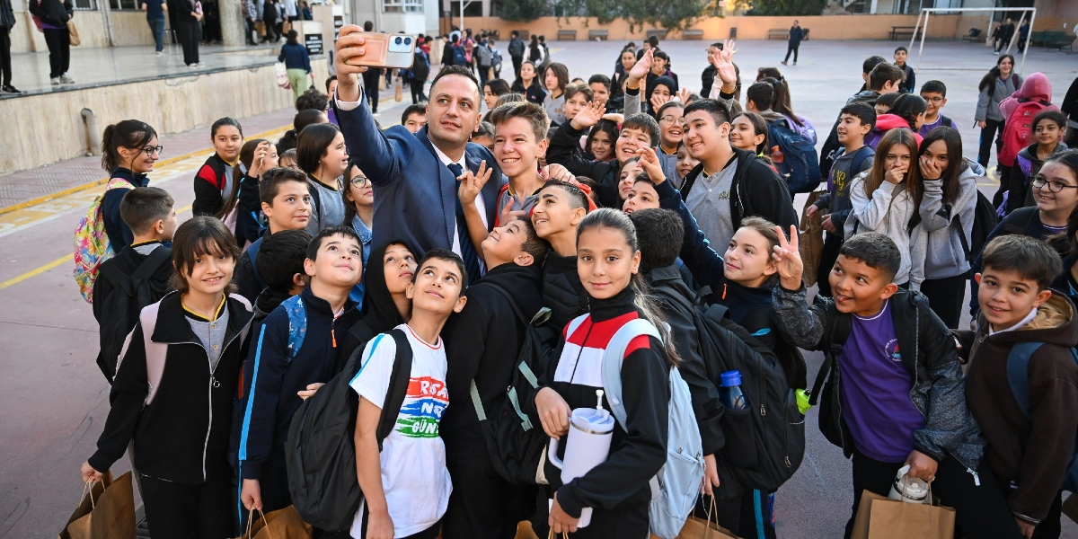 Takım elbiseli bir erkek, okul bahçesinde etrafını saran çok sayıda öğrenciyle birlikte selfie çekiyor. Öğrencilerin birçoğu sırt çantalı ve ellerinde beslenme desteği olduğu anlaşılan kağıt çantalar taşıyor. Bazı öğrenciler gülümseyerek kameraya bakarken, bazıları zafer işareti yapıyor veya arkadaşlarıyla şakalaşıyor. Arka planda okul binasının girişi ve bahçede yürüyen diğer öğrenciler görülüyor.