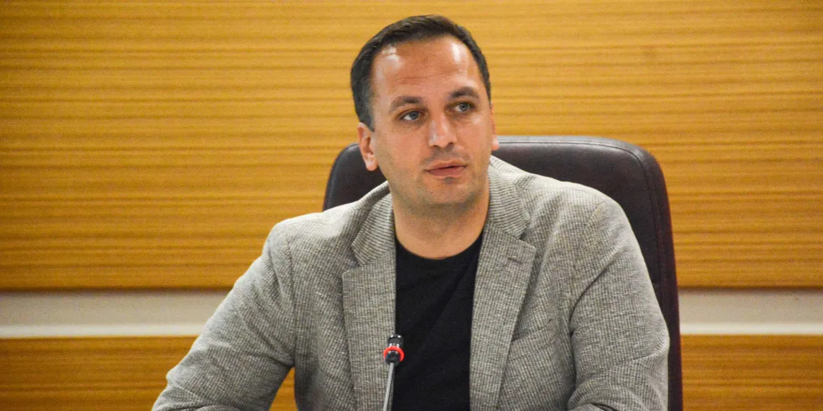 Siyah tişört ve üzerine gri dokulu bir ceket giymiş, kısa saçlı bir erkek, ahşap panellerle kaplı bir duvarın önünde, önünde mikrofon bulunan bir masada oturuyor.