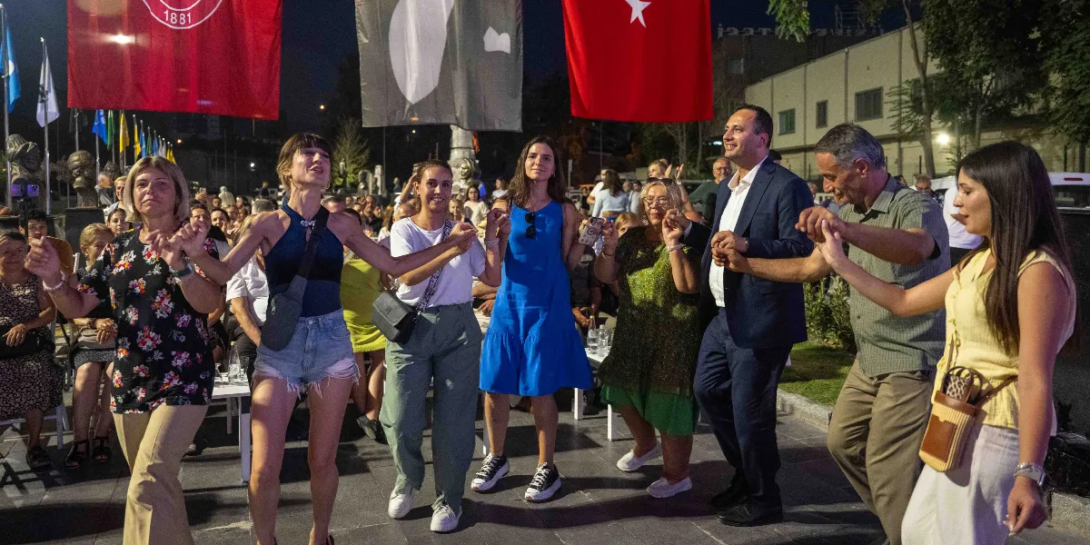 Gece vakti açık havada düzenlenen bir etkinlikte, ellerini birbirine kenetleyerek halay çeken bir grup insan. Arka planda üzerinde 1881 yazılı kırmızı bir bayrak, Türk bayrağı ve diğer flamalar asılı duruyor. Ön planda çiçekli bluz giyen bir kadın, lacivert atletli ve kot şortlu bir genç kadın, beyaz tişörtlü bir kadın, mavi elbiseli bir kadın, yeşil desenli elbise giyen bir kadın, lacivert takım elbiseli bir adam, gri gömlekli bir adam ve sarı bluzlu bir kadın el ele tutuşarak dans ediyor. Arka planda ise etkinliği izleyen kalabalık bir grup yer alıyor.