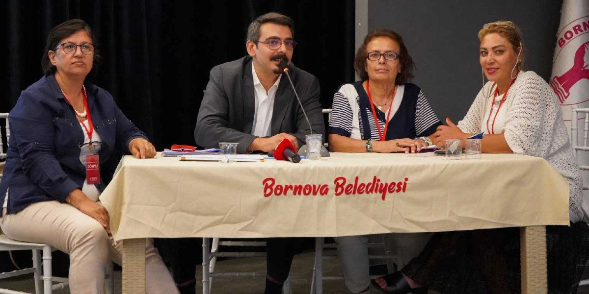 Bornova Belediyesi logolu bir masanın arkasında yan yana oturan dört kişi, bir toplantı veya çalıştay kapsamında katılımcılara hitap ediyor. Soldan sağa doğru; lacivert gömlekli bir kadın, gri ceketli ve gözlüklü bir erkek, çizgili bluzlu bir kadın ve beyaz hırkalı bir kadın yer alıyor. Masanın üzerinde bir mikrofon ve su bardakları bulunuyor, arka planda ise Bornova Belediyesi'nin ambleminin yer aldığı bir bayrak görünüyor.