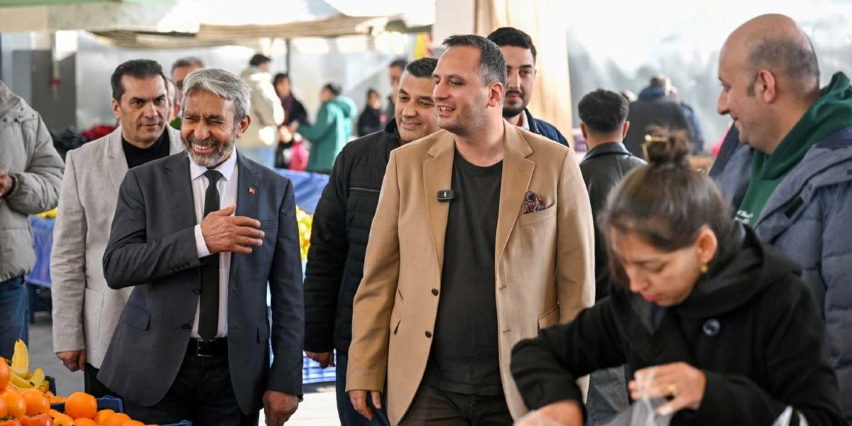 Bej renkli ceket giyen bir erkek, yanında takım elbiseli bir beyefendi ve diğer yetkililerle birlikte İnönü Mahallesi Pazaryeri'nde esnafı ziyaret ediyor. Ön planda bir kadın alışveriş yaparken, arka planda pazar tezgahları ve alışveriş yapan insanlar görülüyor.