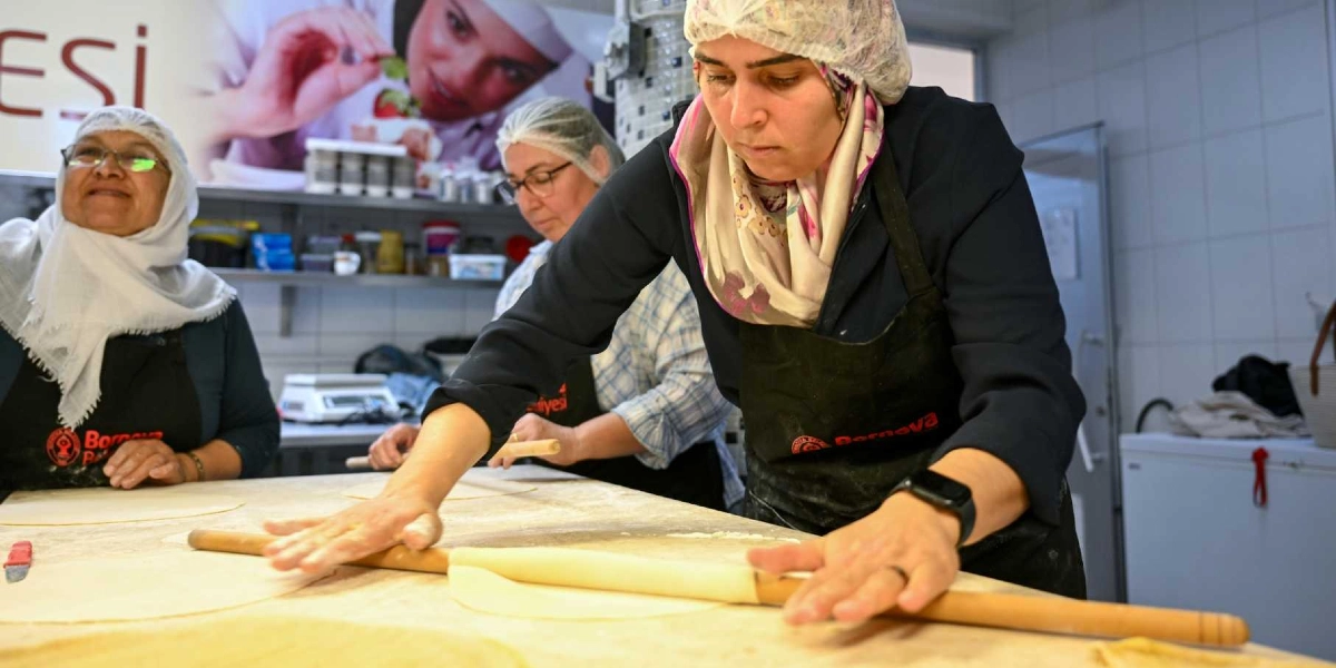 Endüstriyel bir mutfakta, başlarında bone ve üzerlerinde Bornova Belediyesi logolu siyah önlükler bulunan üç kadın, tezgah üzerinde oklava yardımıyla hamur açıyor. Ön planda odak noktasındaki kadın, elleriyle oklavayı kullanarak hamuru inceltirken, arkasında diğer iki kadın da aynı şekilde hamur hazırlığı yapıyor. Arka planda ise üzerinde yemek görseli bulunan bir duvar panosu yer alıyor.