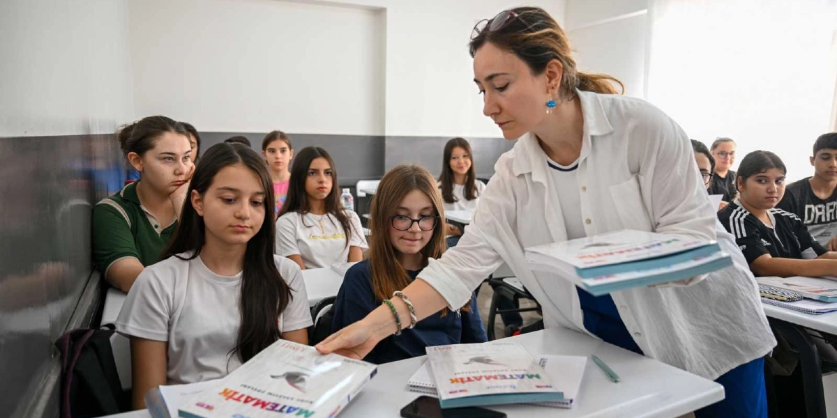 Sınıf ortamında beyaz gömlekli bir kadın öğretmen, öğrencilere üzerinde 