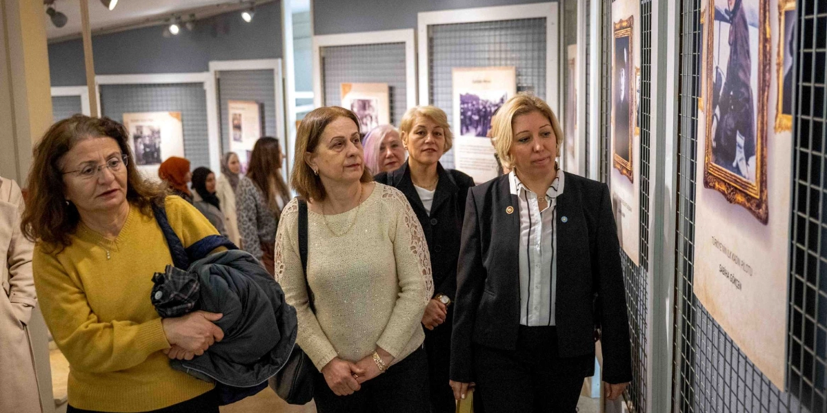 Bir müze sergisinde, duvardaki tarihi fotoğrafları ve bilgilendirme panolarını inceleyen bir grup kadın. Ön planda sarı kazaklı, krem rengi dantel detaylı bluzlu ve siyah ceketli üç kadın, sağ taraftaki duvarda asılı olan ve üzerinde 