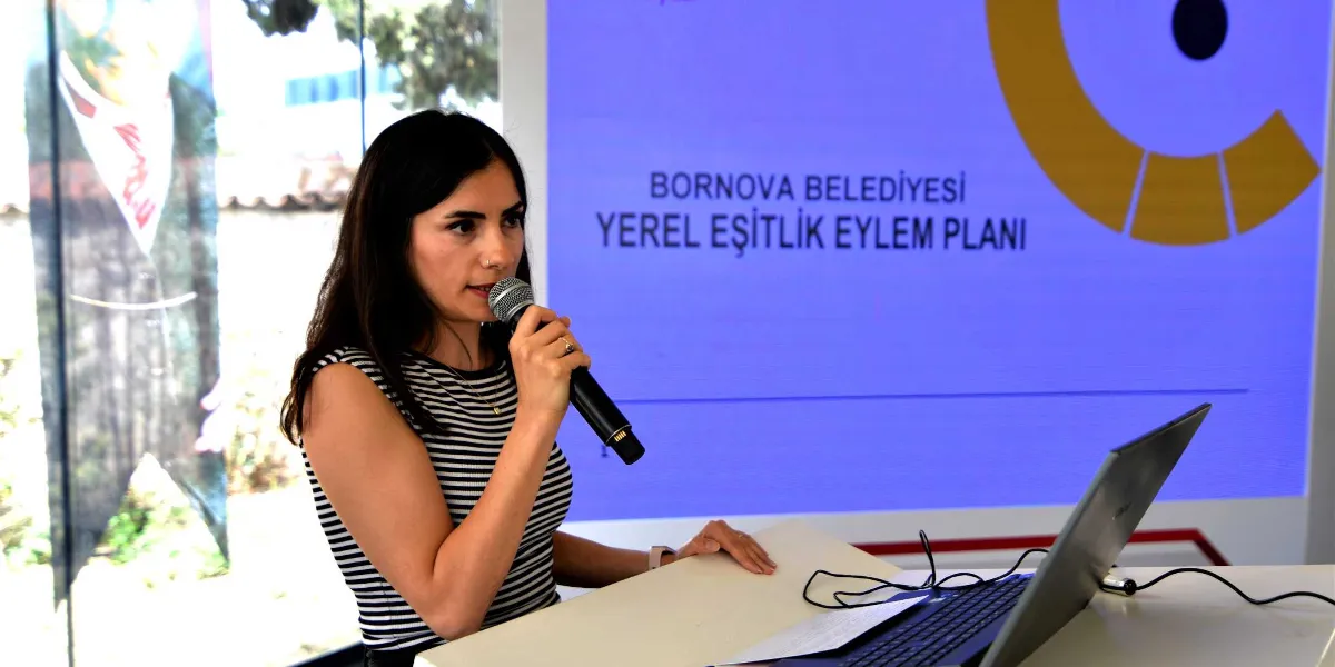 Koyu saçlı, çizgili kolsuz bir bluz giyen bir kadın, kürsüde mikrofonla konuşma yapıyor. Kadının arkasında, üzerinde 