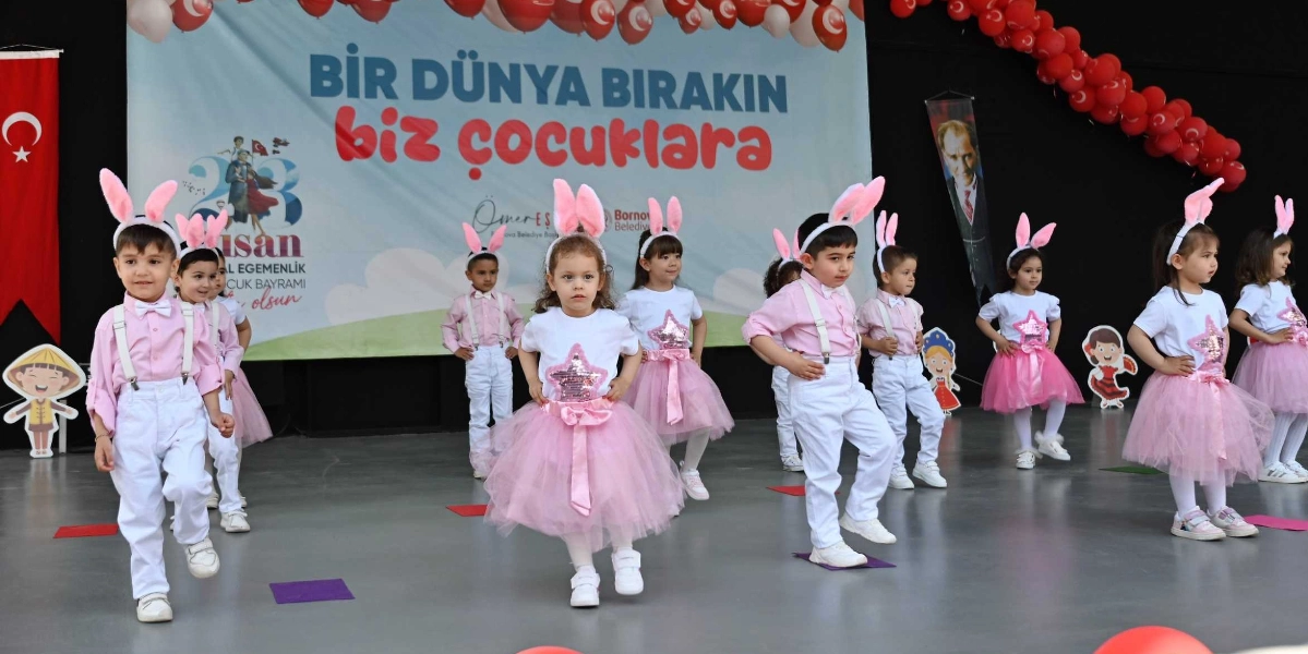 23 Nisan Ulusal Egemenlik ve Çocuk Bayramı kutlamaları kapsamında sahnede dans eden bir grup çocuk. Çocuklar, üzerinde pembe tavşan kulakları olan taçlar takmış; erkek çocuklar pembe gömlek ve beyaz pantolon, kız çocuklar ise beyaz tişört ve pembe tütü etek giymiş. Arka planda 