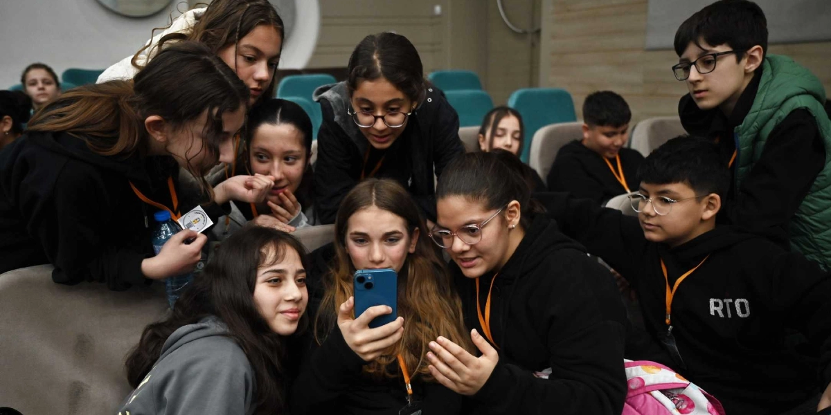 Bir konferans salonunda bir araya gelmiş, boyunlarında turuncu yaka kartları takılı bir grup genç, merkezdeki bir arkadaşının elinde tuttuğu akıllı telefona odaklanmış şekilde bakıyor. Gençler meraklı ve ilgili bir ifadeyle ekranı incelerken, bazıları telefonun etrafında toplanmış, bazıları ise arkadan izliyor. Arka planda salonun koltukları ve diğer katılımcılar seçiliyor.