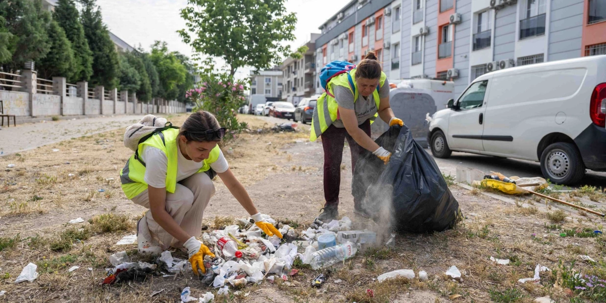 Üzerlerinde sarı ikaz yelekleri ve ellerinde eldivenler bulunan iki kadın, Bornova'da bir sokak kenarındaki çöpleri topluyor. Kadınlardan biri çömelmiş vaziyette yerdeki atıkları eline alırken, diğeri ayakta durarak büyük bir siyah çöp poşetini açık tutuyor. Çevrede binalar, park halindeki araçlar ve ağaçlar yer alıyor.