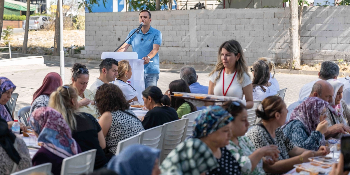 Açık havada düzenlenen bir etkinlikte, mavi polo yaka tişörtlü bir erkek kürsüde mikrofonla konuşma yaparken, önünde oturan kalabalık bir grup yemek yiyor. Beyaz tişörtlü ve boynunda kırmızı bir yaka kartı asılı olan bir kadın, elindeki tepsiyle katılımcılara servis yapıyor. Katılımcıların çoğu kadınlardan oluşuyor ve bir kısmı başörtüsü takıyor. Etkinlik alanı, arkada taş bir duvar ve ağaçların olduğu bir bahçe ortamında gerçekleşiyor.