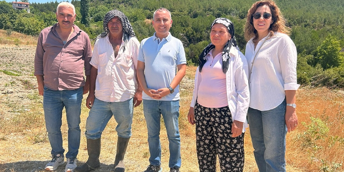 Açık havada, kuru otların bulunduğu bir arazide yan yana duran beş kişilik bir grup. Soldan sağa doğru; kahverengi gömlekli ve kot pantolonlu bir erkek, başında puşi bulunan beyaz gömlekli ve çizmeli bir çiftçi, açık mavi polo yaka tişörtlü ve kot pantolonlu bir erkek, başında çiçekli bir yazma bulunan pembe bluzlu bir kadın ve beyaz gömlekli, güneş gözlüklü bir kadın yer alıyor. Arka planda ağaçlarla kaplı bir yamaç ve uzakta bir ev görülüyor.