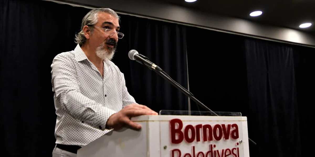 Kır saçlı, gözlüklü ve sakallı bir adam, üzerinde Bornova Belediyesi yazan bir kürsüde mikrofon başında konuşma yapıyor. Adam, beyaz üzerine küçük desenli bir gömlek giymiş ve ellerini kürsünün üzerine koymuş durumda; arka planda ise siyah perdeler yer alıyor.