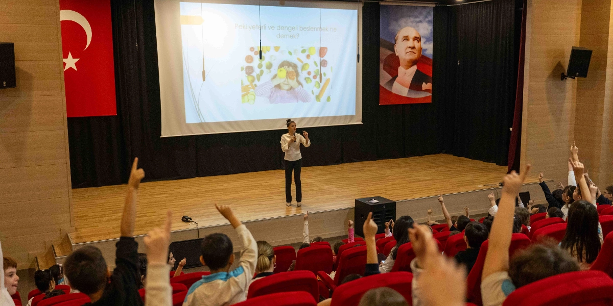Konferans salonunda sahne üzerinde elinde mikrofonla konuşma yapan bir kadın ve onu dinleyen, sorulara cevap vermek için ellerini kaldıran ilkokul çağındaki öğrenciler. Sahnedeki büyük ekranda 