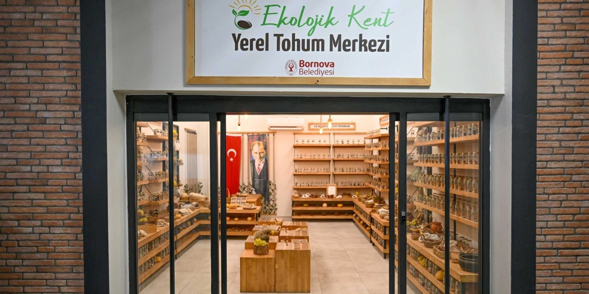 Bornova Belediyesi'ne ait Ekolojik Kent Yerel Tohum Merkezi'nin dış cephesi. Girişin üzerinde merkezin adının yazılı olduğu bir tabela ve altında belediyenin logosu bulunuyor. Cam kapıdan içerisi görünüyor; içeride ahşap raflarda dizili çok sayıda tohum kavanozu, ortada ahşap stantlar, duvarda asılı bir Türk bayrağı ve Mustafa Kemal Atatürk'ün portresi yer alıyor. Merkezin dış duvarları kırmızı tuğla kaplı.