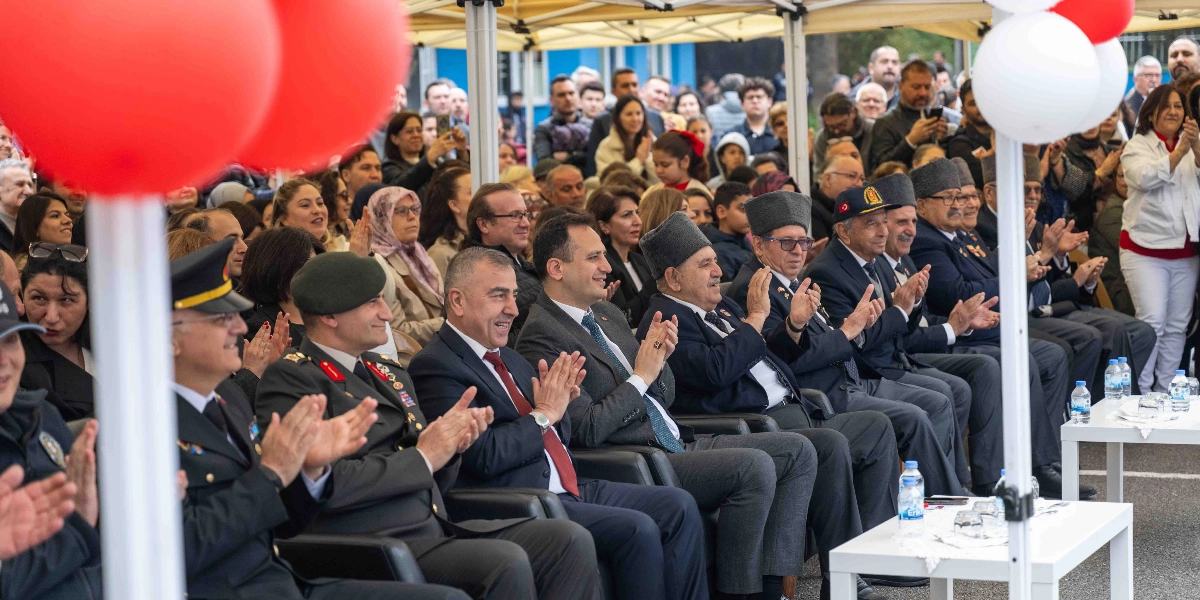 Açık havada düzenlenen bir etkinlikte, protokol üyeleri ve vatandaşlar alkışlayarak bir gösteriyi izliyor. Ön sırada askeri üniformalı yetkililer, takım elbiseli yöneticiler ve geleneksel kalpak giymiş yaşlı beyefendiler yan yana oturuyor. Arka planda ise etkinliği takip eden kalabalık bir grup izleyici yer alıyor. Etkinlik alanı kırmızı ve beyaz balonlarla süslenmiş durumda.