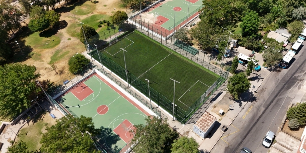 Yeşil alan içerisinde yer alan, tel örgülerle çevrili dikdörtgen bir halı saha ve hemen yanında bulunan basketbol sahasının kuş bakışı görünümü. Sahaların çevresinde ağaçlar, küçük bir yapı ve sağ tarafta araçların geçtiği bir yol bulunmaktadır.