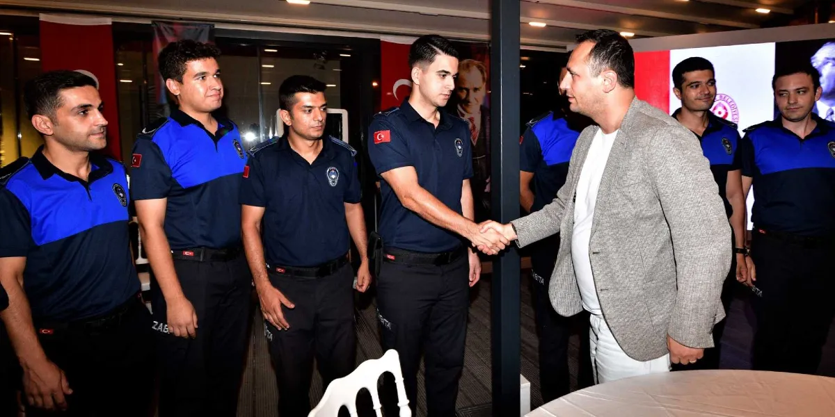 Belediye başkanı, Zabıta Haftası kapsamında düzenlenen bir etkinlikte, mavi ve lacivert üniformalı zabıta memurlarıyla bir araya gelerek bir memurun elini sıkıyor. Arka planda Türk bayrağı ve Atatürk portresi yer alıyor.
