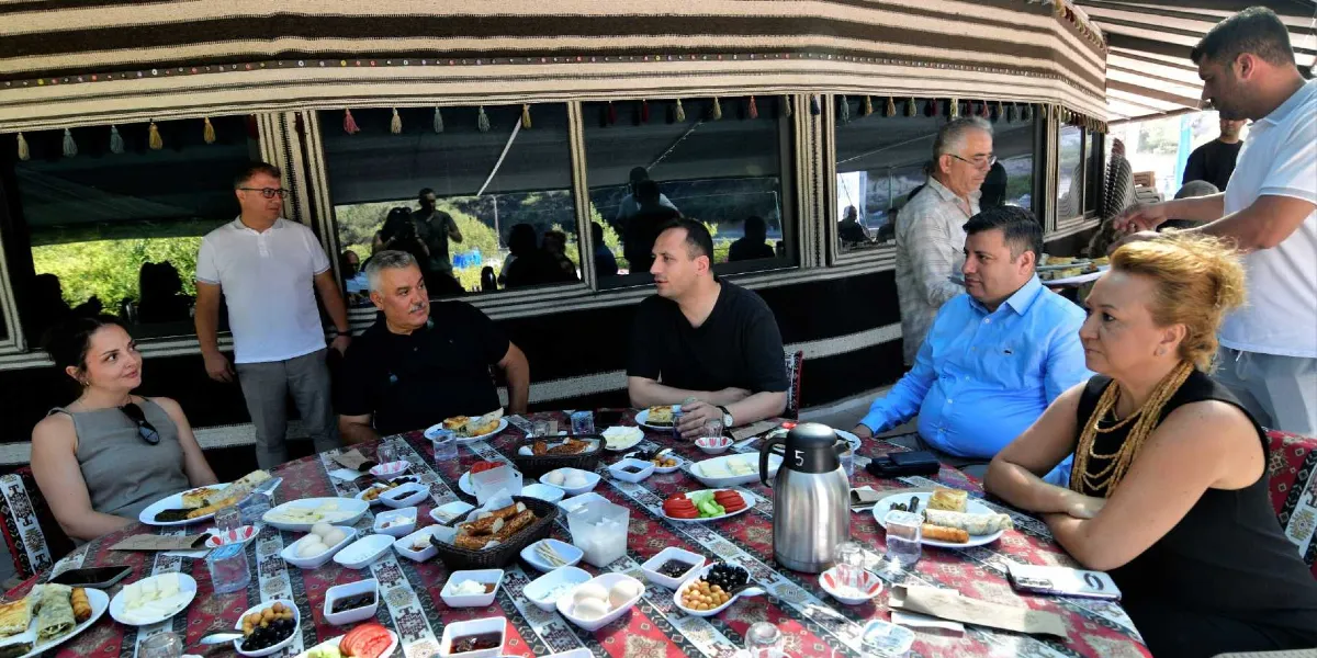 Geleneksel desenli bir Yörük çadırının içinde, uzun bir masanın etrafında toplanmış bir grup insan kahvaltı yapıyor. Masada çeşitli peynirler, zeytinler, domatesler, börekler ve çay servisleri bulunuyor. Çadırın duvarları geleneksel dokuma kumaşlarla kaplı ve pencerelerden dışarıdaki yeşillik görünüyor. Masanın başında ve çevresinde oturan farklı yaş ve giyim tarzındaki kişiler sohbet ederek yemeklerini yiyor, ayakta duran bir kişi ise servis yapmaya hazırlanıyor.