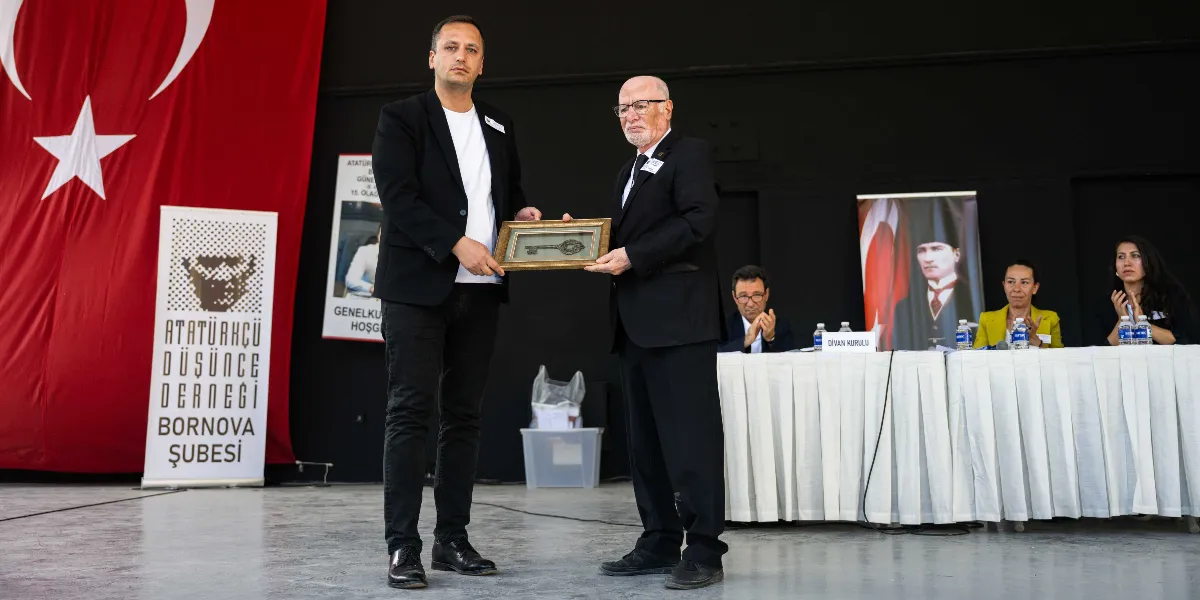 Atatürkçü Düşünce Derneği Bornova Şubesi genel kurulunda, siyah ceketli iki erkek, çerçeve içinde eski bir anahtar figürünü birbirlerine takdim ediyor. Arka planda büyük bir Türk bayrağı, Atatürkçü Düşünce Derneği Bornova Şubesi yazılı bir dikey pankart ve üzerinde Atatürk portresi bulunan bir başka pankart yer alıyor. Sağ tarafta, beyaz örtülü bir masada oturan divan kurulu üyeleri alkışlayarak törene eşlik ediyor.