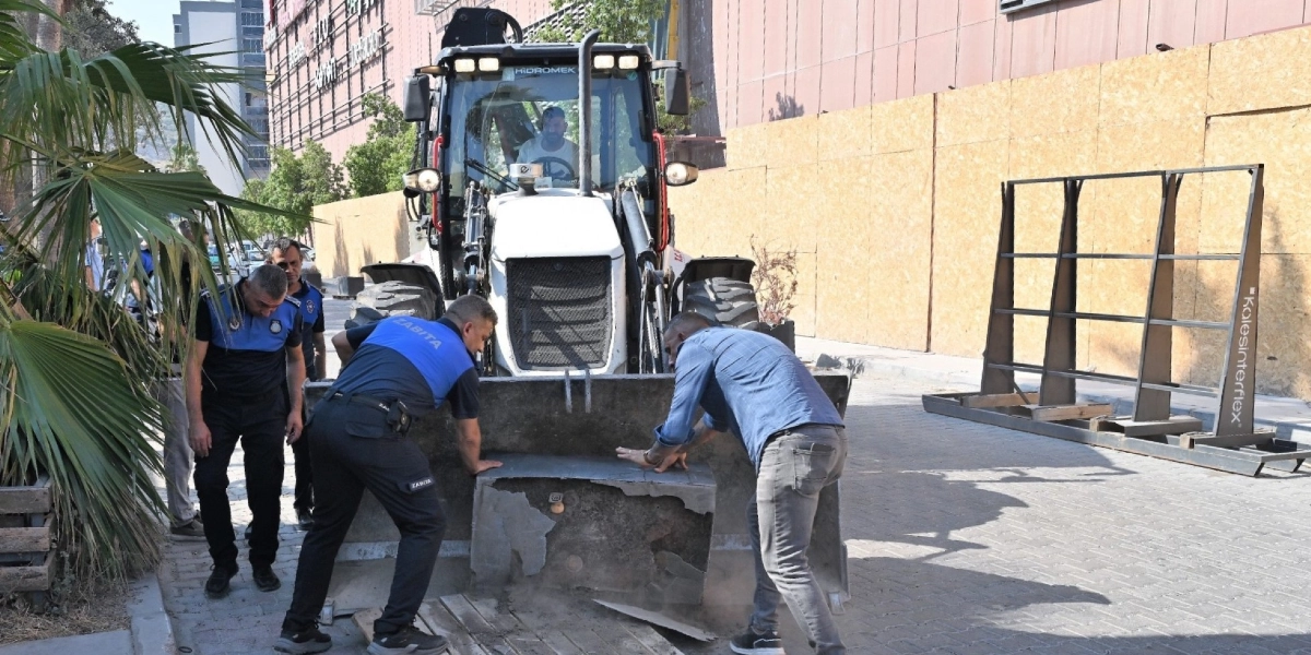 Bornova'da zabıta ekipleri, kaldırım işgaline neden olan beton bir bloğu iş makinesinin kepçesine yükleyerek kaldırmaya çalışıyor. Ekiplerin yanında sivil giyimli bir kişi de sürece yardımcı oluyor.
