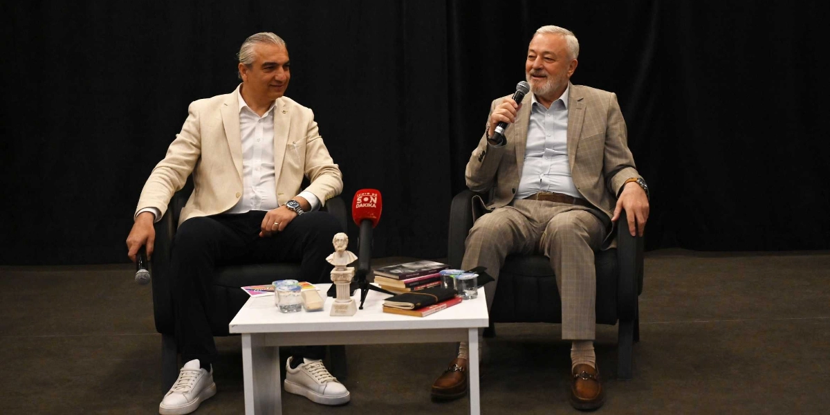 Siyah bir fon önünde, beyaz bir sehpanın iki yanında oturan iki erkek konuşmacı. Sağ tarafta oturan, ekose ceketli ve gri saçlı kişi elinde mikrofonla konuşurken, sol tarafta oturan, bej ceketli ve beyaz spor ayakkabılı kişi onu dinliyor. Sehpanın üzerinde küçük bir büst, birkaç kitap, su bardakları ve üzerinde 