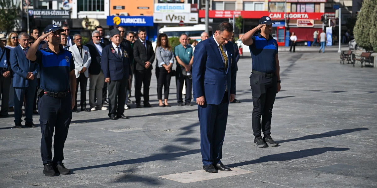 Bornova'daki Gaziler Günü töreninde, lacivert takım elbiseli bir yetkili başı öne eğik şekilde saygı duruşunda bulunurken, iki yanında üniformalı polis memurları selam duruşu yapıyor. Arka planda ise resmi kıyafetli gaziler ve sivil katılımcılar töreni takip ediyor.