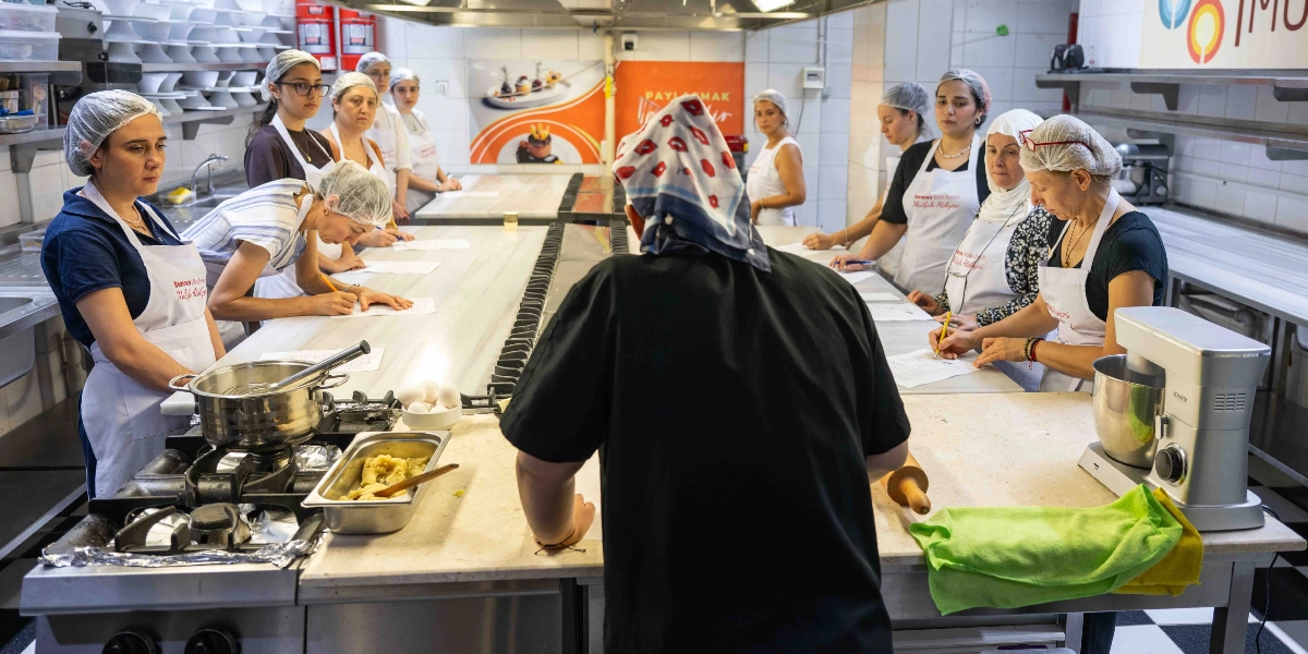 Profesyonel bir mutfak ortamında, başlarında bone takan kadın kursiyerlerin katıldığı bir yemek atölyesi. Uzun mermer tezgâhların etrafında toplanan katılımcılar, önlerinde bulunan tarif kâğıtlarına notlar alıyor. Ön planda siyah üniformalı bir eğitmen, tezgâhın başında durarak süreci yönetiyor. Mutfak tezgâhlarının üzerinde paslanmaz çelik tencereler, yumurtalar, oklava ve bir mutfak şefi mikseri bulunuyor. Arka planda ise Bornova Belediyesi Mutfak Atölyesi'ne ait logolar ve mutfak ekipmanları yer alıyor.