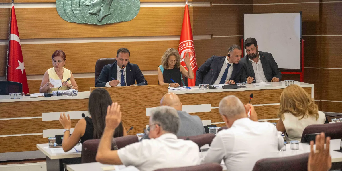 Bornova Belediyesi meclis salonunda düzenlenen toplantıda, katılımcılar ellerini kaldırarak oylama yapıyor. Masanın ön tarafında oturan meclis üyeleri ve arka tarafta kürsüde yer alan yetkililer, önlerindeki evrakları inceliyor. Salonun arka duvarında Türk bayrağı ve Bornova Belediyesi'nin logosunun bulunduğu bayrak asılı duruyor.