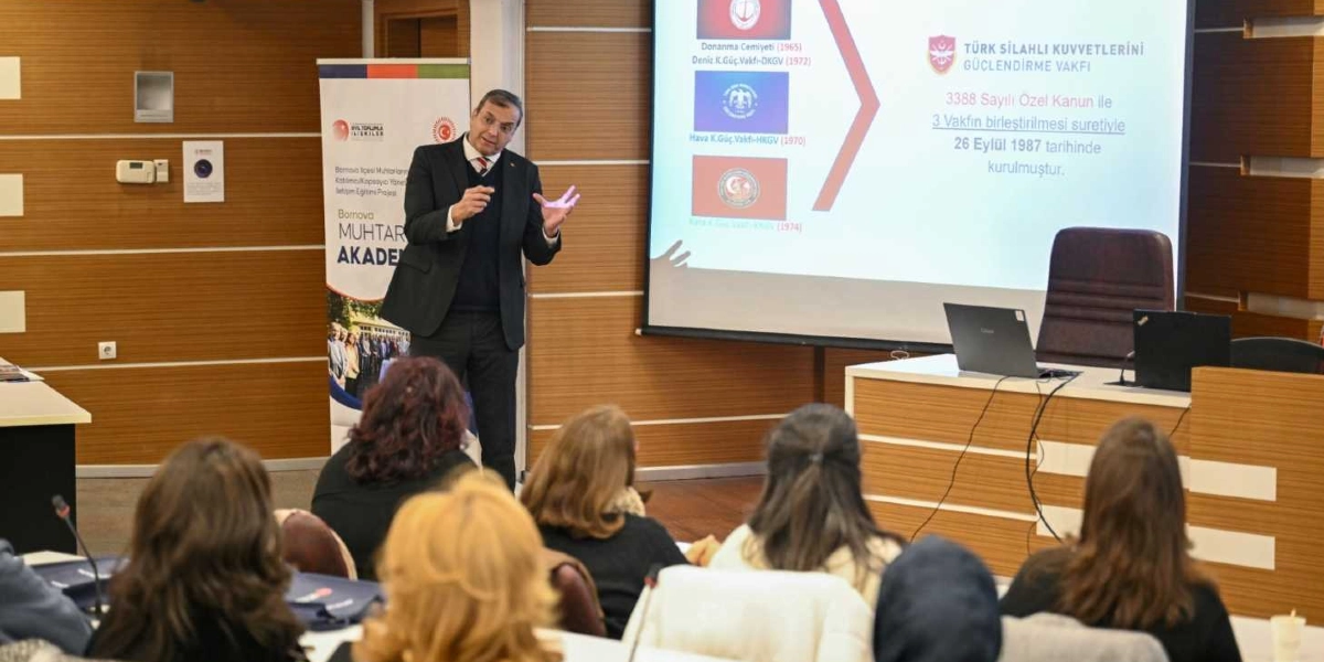 Modern bir toplantı salonunda, Bornova Muhtar Akademisi kapsamında bir sunum yapan konuşmacı ve onu dinleyen katılımcılar yer alıyor. Siyah takım elbiseli konuşmacı, ayakta durarak elleriyle bir noktayı işaret ediyor. Arkasındaki projeksiyon perdesinde Türk Silahlı Kuvvetlerini Güçlendirme Vakfı'nın tarihçesi, üç vakfın birleşimi ve 26 Eylül 1987 kuruluş tarihi bilgileri yer alıyor. Salonun ön tarafında, üzerinde dizüstü bilgisayar bulunan bir kürsü ve yanında Bornova Muhtar Akademisi'ni tanıtan dikey bir afiş bulunuyor. Katılımcılar ise arkaları dönük şekilde sıralarda oturarak sunumu takip ediyor.