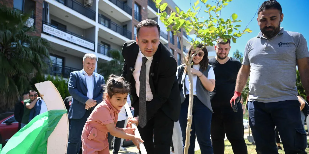Bornova Belediyesi tarafından düzenlenen bir etkinlikte, takım elbiseli bir yetkili ve küçük bir kız çocuğu birlikte fidan dikiyor. Çevredeki diğer katılımcılar ve belediye personeli gülümseyerek bu anı izliyor. Arka planda ise üzerinde 