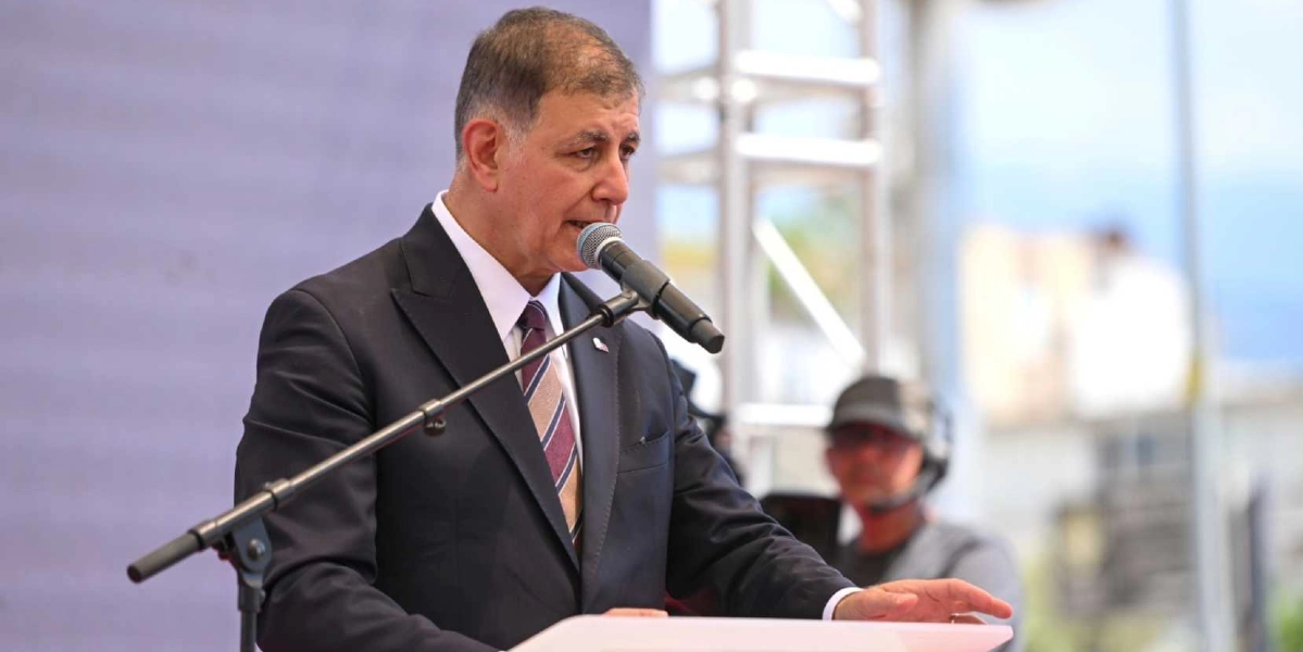 Koyu renk takım elbise, beyaz gömlek ve çizgili kravat giymiş bir erkek, kürsüde mikrofon başında konuşma yapıyor. Arka planda ise bulanık bir şekilde bir kameraman görülüyor.