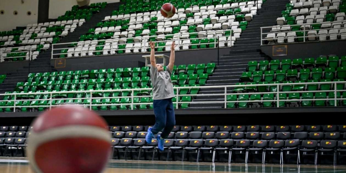 Gri tişört ve koyu renk pantolon giyen genç bir basketbolcu, boş tribünlerin önünde havaya sıçrayarak atış yaparken; ön planda ise odak dışı kalmış bir basketbol topu görülüyor.