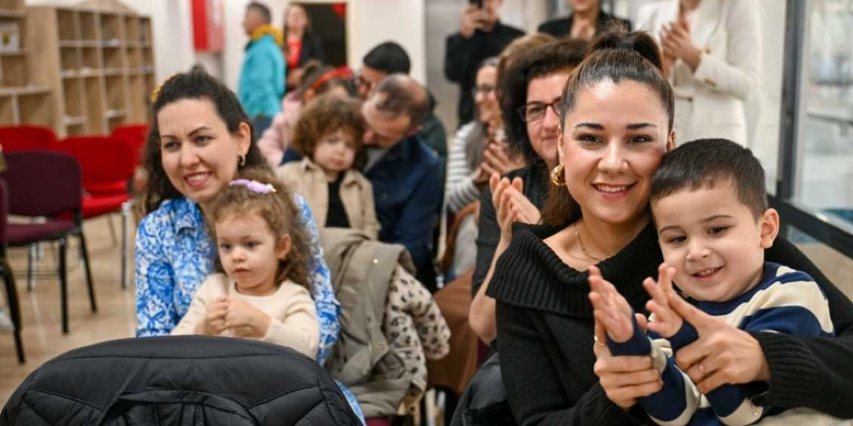 Kapalı bir mekanda, bir etkinlikte yan yana oturan ve gülümseyerek alkışlayan ebeveynler ve çocukları. Ön planda siyah kazaklı bir kadın, kucağındaki küçük erkek çocuğunun ellerini tutarak alkışlamasına yardımcı oluyor. Yanlarında mavi desenli kıyafetli bir kadın ve kucağında küçük bir kız çocuğu oturuyor. Arka planda ise etkinliği izleyen diğer katılımcılar yer alıyor.