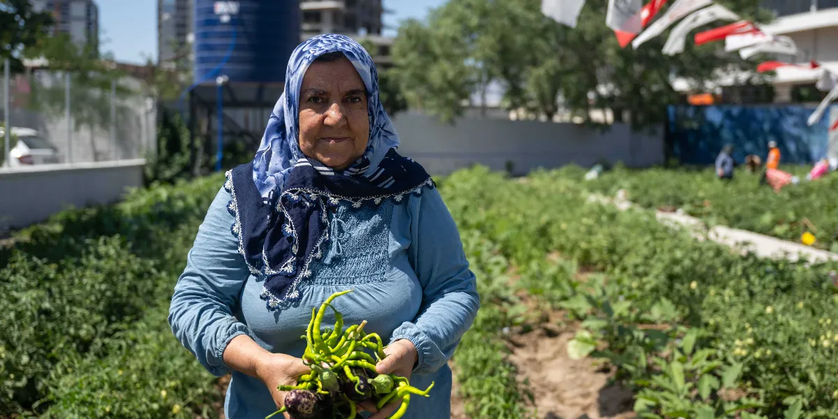 Mavi başörtülü ve açık mavi bluzlu orta yaşlı bir kadın, ellerinde yeni topladığı yeşil biberler ve patlıcanlarla bir sebze bahçesinde duruyor. Arka planda yeşil bitkilerle dolu bahçe ve uzakta bazı yapılar görülüyor.
