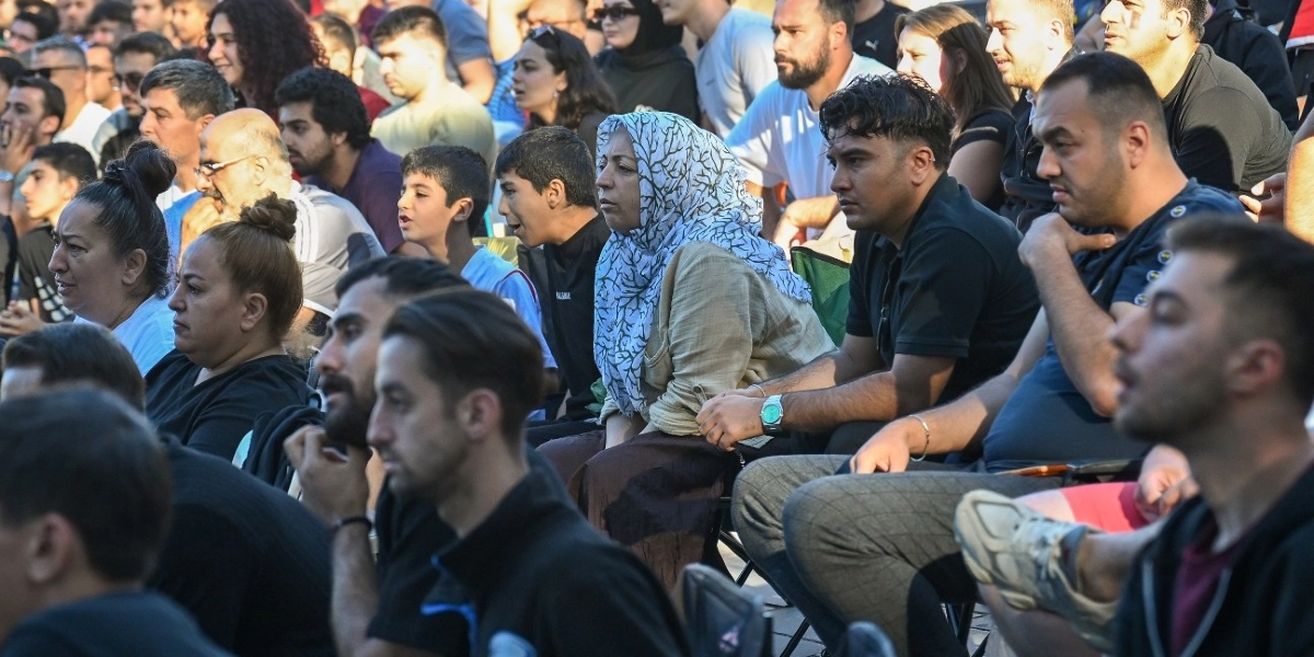 Açık havada bir etkinlikte yan yana oturmuş, dikkatle bir yöne doğru bakan, farklı yaş ve cinsiyetlerden oluşan kalabalık bir grup insan. Ön planda, beyaz üzerine siyah ağaç dalı desenli bir başörtüsü takan bir kadın ve yanında koyu renkli tişörtler giymiş genç erkekler yer alıyor. Arka planda ise daha fazla izleyici bulunuyor ve ortamda genel bir topluluk havası hakim.