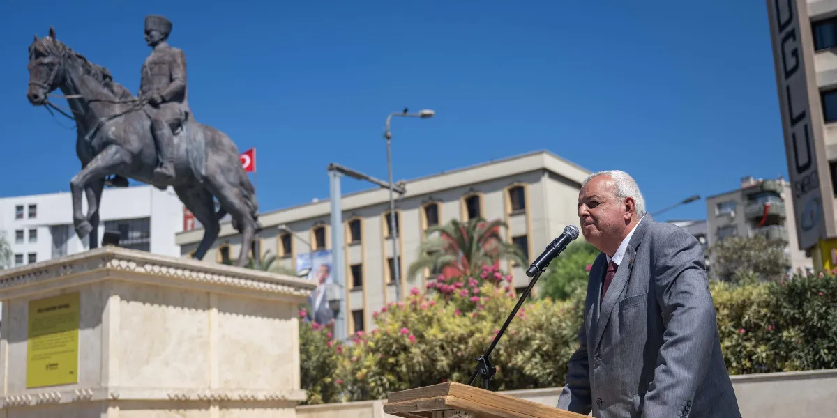 Açık havada, arkasında Mustafa Kemal Atatürk'ün at üzerindeki heykelinin bulunduğu bir kürsüde konuşma yapan gri takım elbiseli ve kravatlı bir erkek. Arka planda binalar, çiçekli bitkiler ve gökyüzünde dalgalanan Türk bayrağı görülüyor.