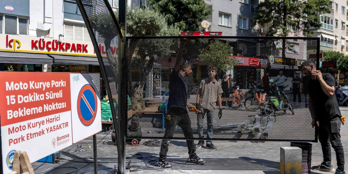Bornova'da bir sokakta, üç işçi metal bir gölgelik panelinin montajını yapıyor. İşçilerden biri elindeki su terazisiyle panelin dengesini kontrol ederken, diğerleri montaj sürecine destek veriyor. Yan tarafta, üzerinde 