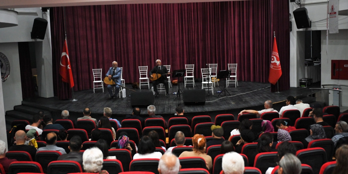 Kırmızı perdeli bir sahne üzerinde, ellerinde bağlama tutan iki müzisyen sandalyelerde oturarak performans sergiliyor. Sahnenin iki yanında Türk bayrağı ve Bornova Belediyesi'nin flaması yer alıyor. Sahne önündeki izleyici koltukları dolu olup, dinleyiciler sahnedeki müzik dinletisini takip ediyor.