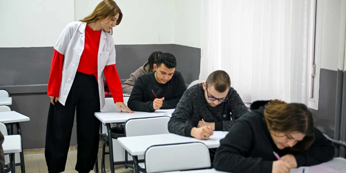 Sınıf ortamında deneme sınavına giren öğrenciler ve onları gözlemleyen bir öğretmen. Kırmızı kazak ve beyaz önlük giymiş kadın öğretmen, sıraların arasında yürürken öğrencileri kontrol ediyor. Öğrenciler ise sıralarında oturmuş, odaklanmış bir şekilde sınav kağıtlarına yazı yazıyor.