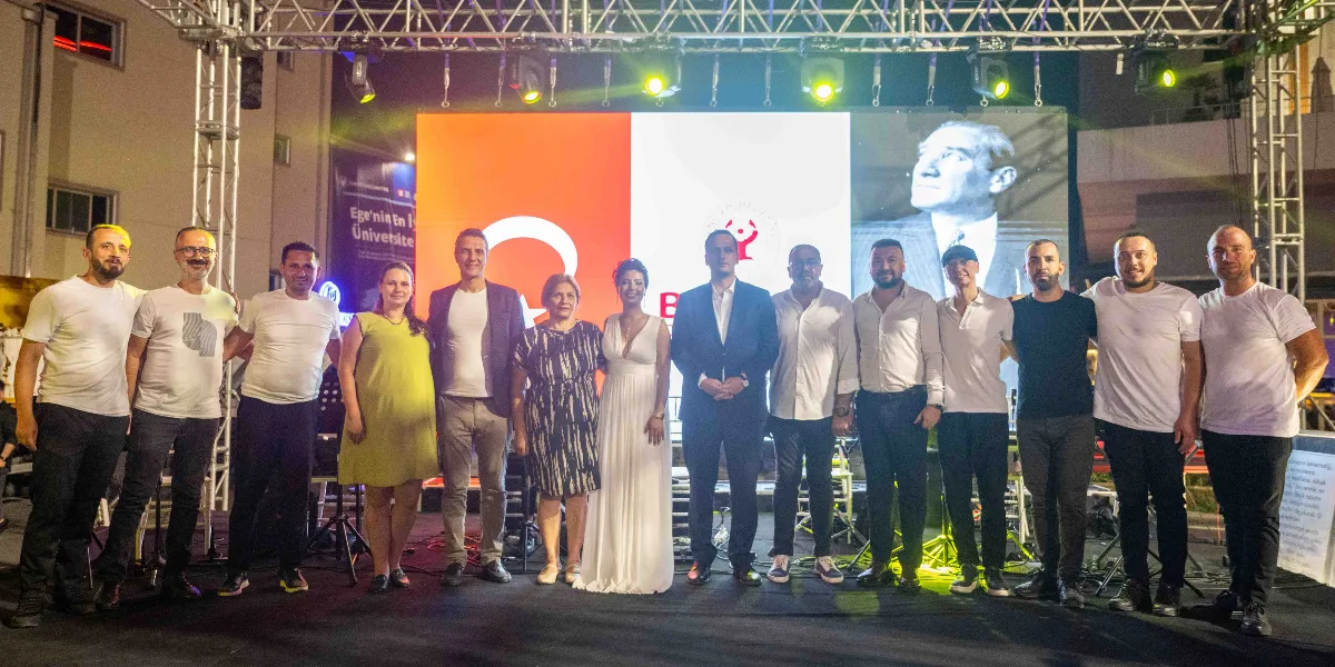 Gece vakti açık hava sahnesinde yan yana dizilmiş, çoğu beyaz tişörtlü olan on dört kişilik bir grup, arkalarındaki dev ekranda Mustafa Kemal Atatürk'ün silüeti ve Bornova Belediyesi logosunun önünde poz veriyor. Sahne ışıklarıyla aydınlatılmış grupta, beyaz uzun bir elbise giyen bir kadın ve takım elbiseli bir erkek de yer alıyor.