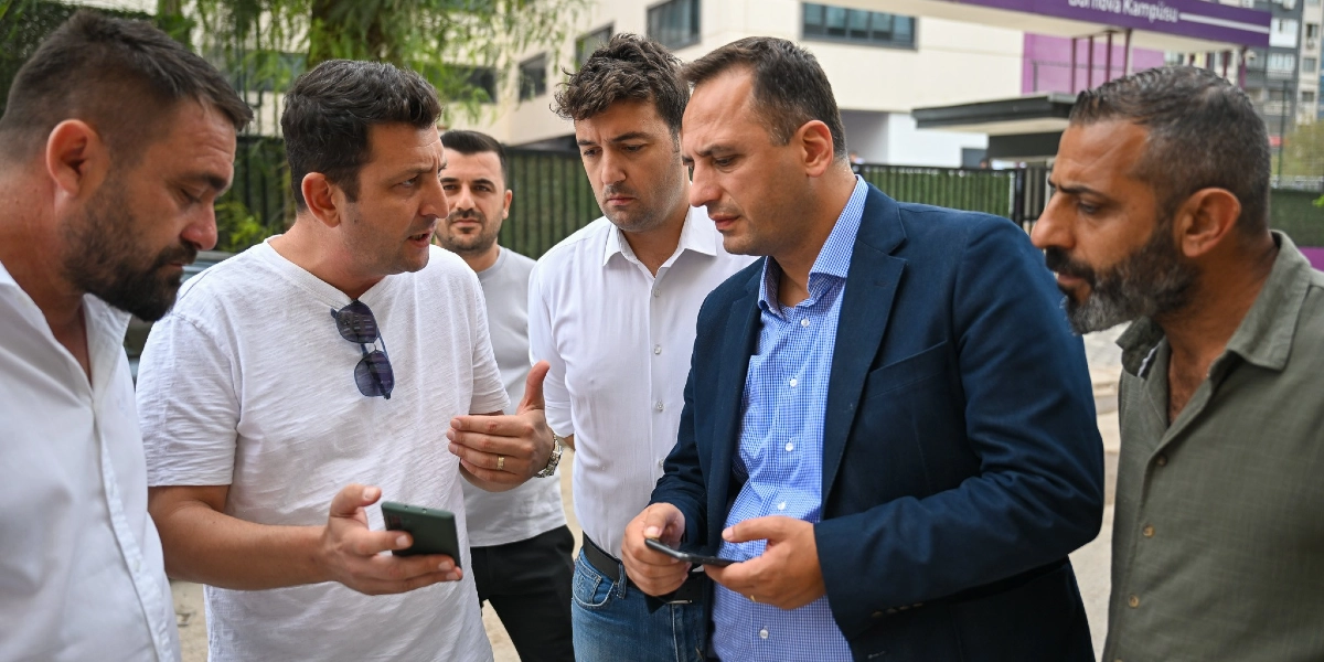 Bornova'da bir cadde üzerinde bir araya gelen beş kişilik bir grup erkek, ellerindeki telefonlara bakarak ve birbirleriyle konuşarak bir proje veya çalışma hakkında fikir alışverişinde bulunuyor. Gruptakilerden biri beyaz tişörtlü, diğeri ise lacivert ceketli ve mavi gömleklidir; diğerleri ise günlük kıyafetler içindedir. Arka planda Bornova Kampüsü yazılı bir tabela ve ağaçlık bir alan görülmektedir.