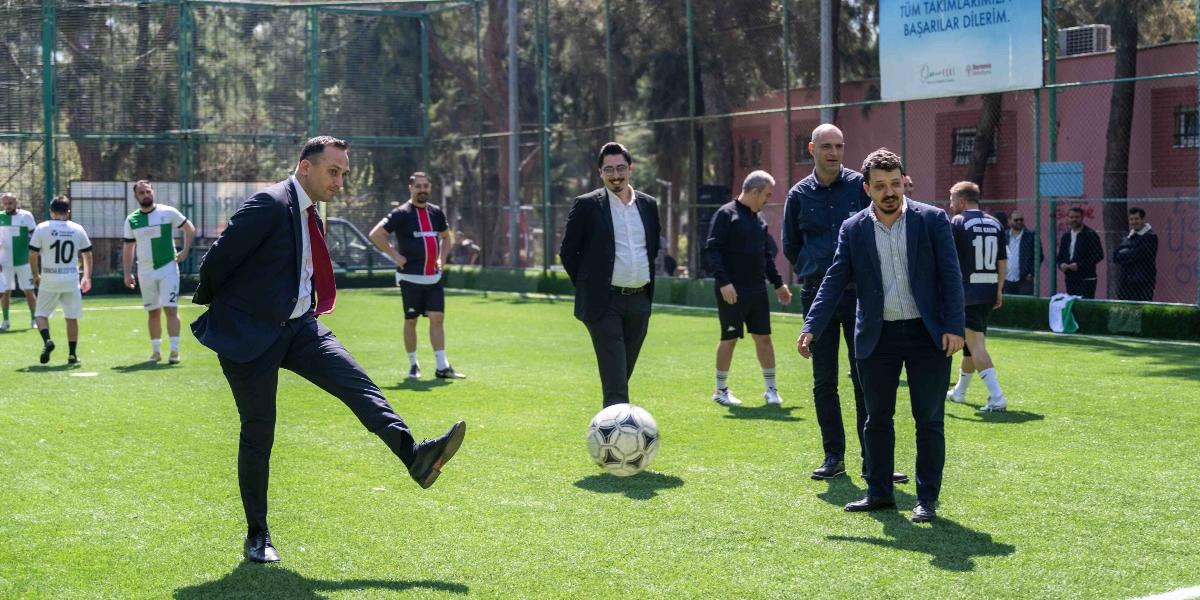 Lacivert takım elbiseli bir erkek, halı sahada futbol topuna vurarak turnuvanın başlama vuruşunu yapıyor. Çevresinde spor kıyafetli oyuncular ve takım elbiseli izleyiciler bulunuyor. Arka planda ise üzerinde 
