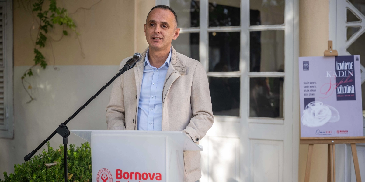 Açık renkli ceket giymiş bir erkek, Bornova Belediyesi logolu bir kürsüde mikrofon başında konuşma yapıyor. Arka planda, üzerinde 
