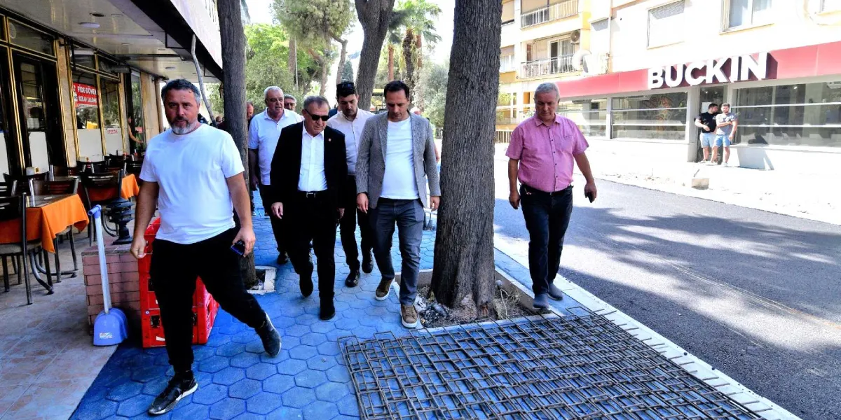 Beş erkekten oluşan bir grup, yenilenmekte olan bir sokakta yürüyerek incelemelerde bulunuyor. Grubun önünde beyaz tişörtlü bir adam, arkasında ise takım elbiseli ve günlük kıyafetli diğer yetkililer yer alıyor. Zemin, mavi altıgen taşlarla döşenmiş olup, yer yer inşaat çalışmaları için kullanılan metal hasır donatılar görülüyor. Arka planda ise 