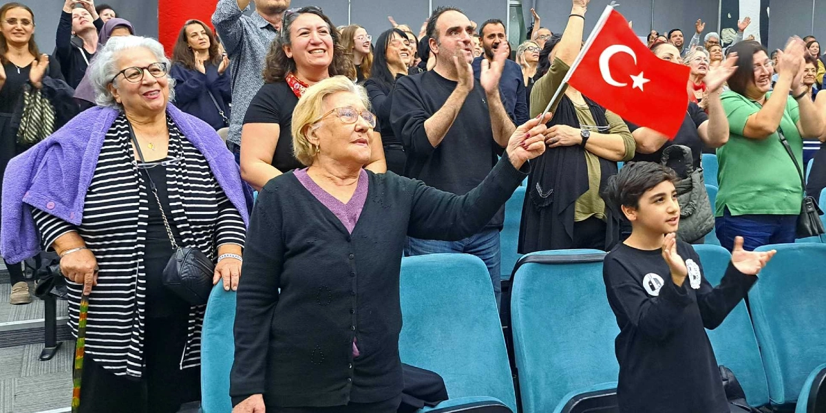 Kapalı bir salonda, mavi koltuklarda oturan veya ayakta duran bir grup insan, coşkuyla alkışlıyor. Ön planda, sarı saçlı, gözlüklü bir kadın elinde Türk bayrağı tutuyor. Yanında siyah tişörtlü bir çocuk ve sol tarafta çizgili bluzunun üzerine mor bir hırka giymiş, elinde baston tutan başka bir kadın yer alıyor. Arka planda ise farklı yaş gruplarından insanlar, ellerini havaya kaldırarak alkışlıyor ve gülümseyerek sahneye bakıyor.