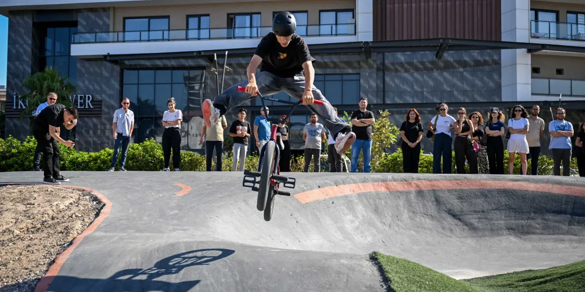 Siyah kasklı ve tişörtlü bir genç, BMX bisikletiyle beton bir parkurda havaya sıçrayarak akrobasi hareketi yapıyor. Arka planda modern bir bina ve bu gösteriyi ilgiyle izleyen bir grup insan bulunuyor.