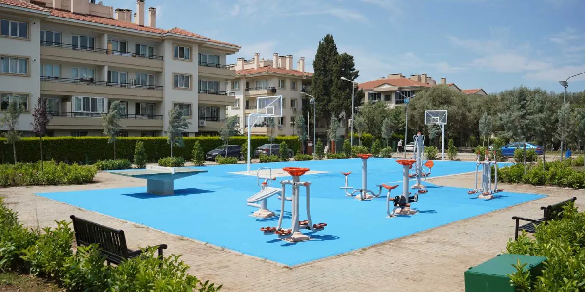 Mavi zeminli bir açık hava spor ve oyun alanında, çeşitli dış mekan egzersiz aletleri, bir masa tenisi masası ve arka planda basketbol potaları bulunuyor. Parkın çevresi yeşil bitkilerle çevrili olup, arka planda çok katlı konut binaları ve ağaçlar yer alıyor.
