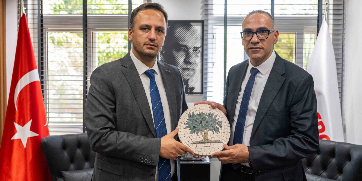 Takım elbiseli iki erkek, ellerinde üzerinde zeytin ağacı figürü bulunan dairesel bir mozaik tabloyu birlikte tutuyor. Arka planda bir yanda Türk bayrağı, diğer yanda ise kısmen görünen beyaz bir bayrak yer alıyor. İkilinin arkasındaki duvarda ise siyah beyaz bir portre tablosu bulunuyor.