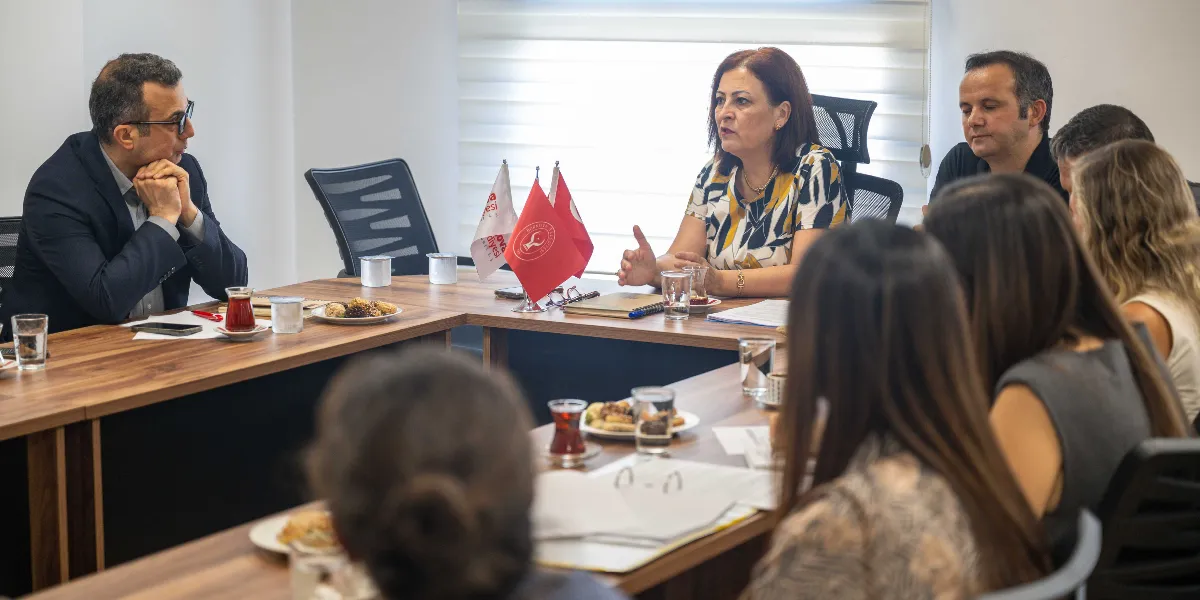 U-şeklindeki bir toplantı masasında bir araya gelen bir grup insan, Bornova Belediyesi'ne ait bayrakların bulunduğu masada görüşme yapıyor. Masanın başında oturan kadın konuşurken, karşısında oturan gözlüklü bir erkek onu dinliyor; masada çay bardakları ve atıştırmalık tabakları yer alıyor.
